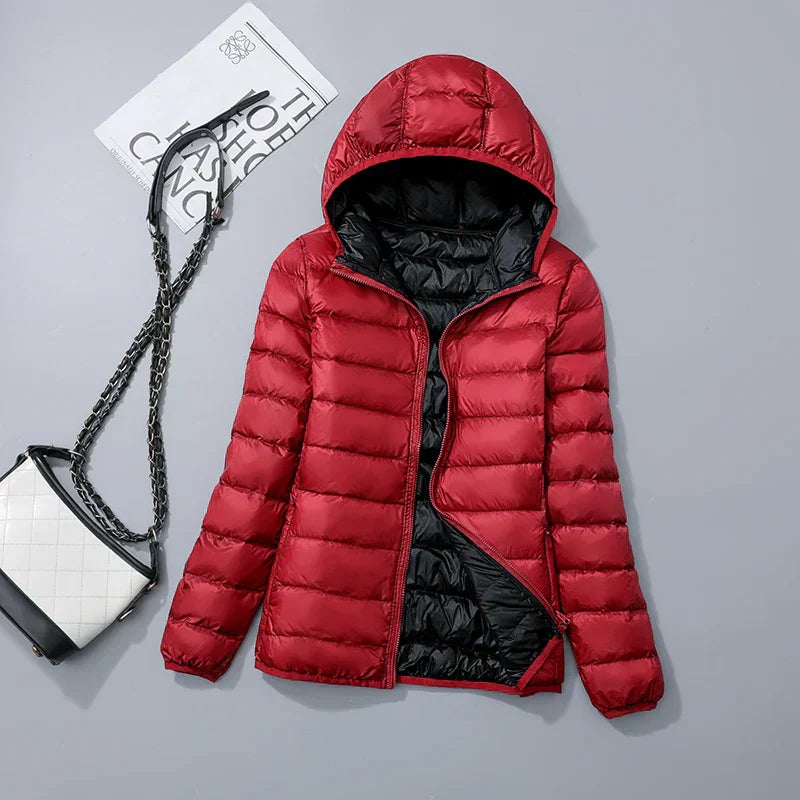 Veste doudoune rouge avec capuche, posée à plat, accompagnée d'un sac à main noir et blanc à chaîne, sur fond gris. Mode hiver tendance.