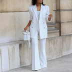 Femme en costume blanc élégant, blazer à boutons dorés, pantalon évasé, sac à main assorti, posant devant un mur en pierre. Mode chic et moderne.