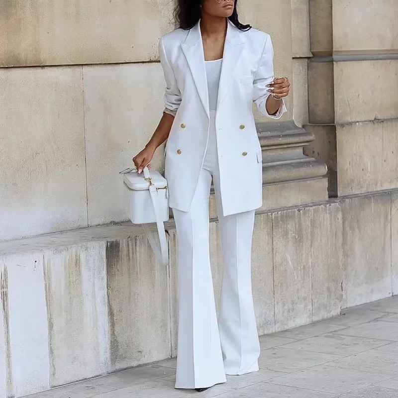 Femme en costume blanc élégant, blazer à boutons dorés, pantalon évasé, sac à main assorti, posant devant un mur en pierre. Mode chic et moderne.