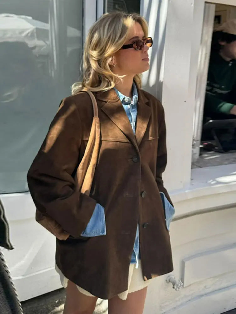 Femme blonde portant un manteau marron, chemise en jean et lunettes de soleil, debout devant une vitrine. Mode automne, style urbain chic.
