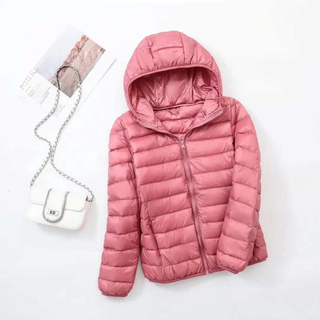 Veste matelassée rose avec capuche, posée à côté d'un sac à main blanc à chaîne argentée sur fond blanc. Mode hiver femme tendance.