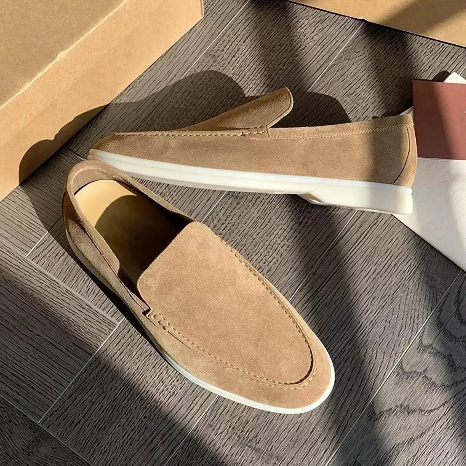 Mocassins en daim beige pour homme sur parquet en bois, éclairés par le soleil. Chaussures élégantes avec semelle blanche, style décontracté chic.