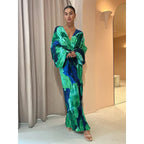 Robe longue plissée verte et bleue, manches larges, portée par une femme élégante dans un intérieur moderne. Mode féminine, style chic.