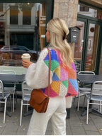 Femme blonde portant un pull coloré en patchwork, tenant un café à emporter, marchant devant un café avec des chaises métalliques à l'extérieur.