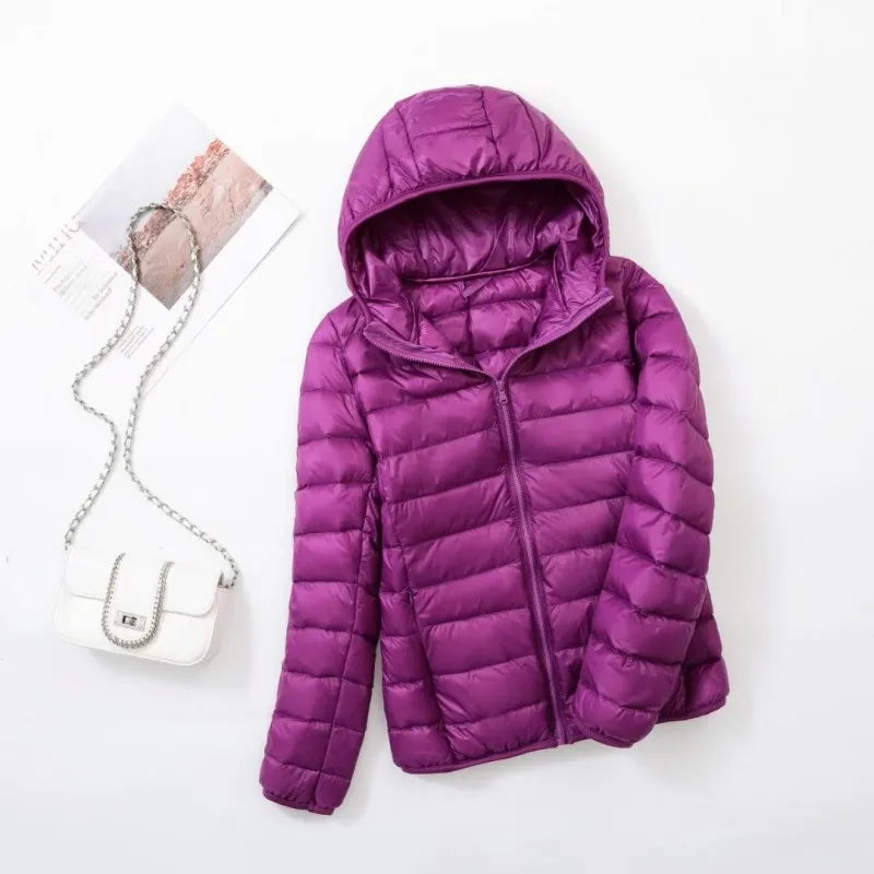 Veste doudoune violette avec capuche, posée à plat, accompagnée d'un sac à main blanc à chaîne argentée et d'un magazine sur fond blanc.