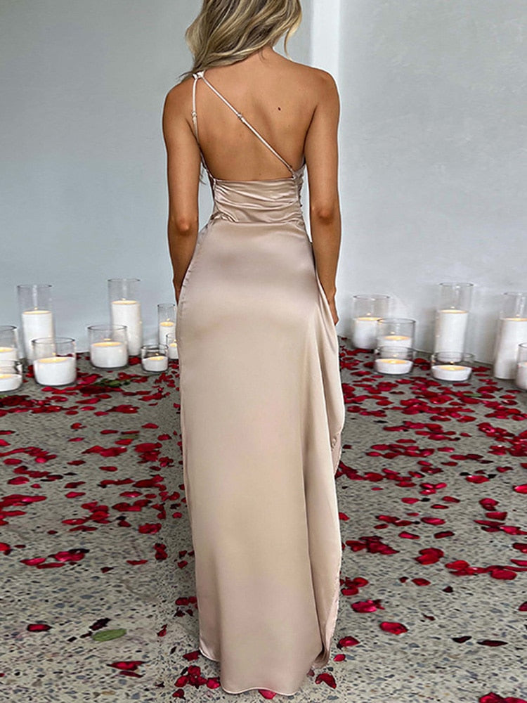 Robe de soirée longue en satin beige, dos nu, avec bretelle fine, entourée de pétales de rose rouges et bougies blanches sur sol en pierre.