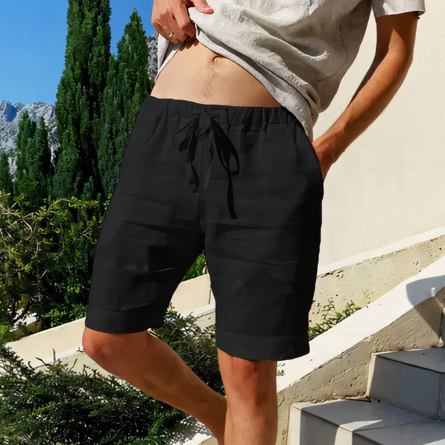 Homme en short noir sur une terrasse ensoleillée, entouré de verdure et montagnes en arrière-plan, portant un t-shirt beige. Mode estivale décontractée.