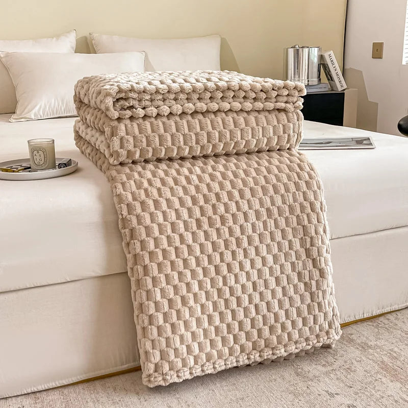 Couverture en laine beige texturée sur un lit blanc dans une chambre moderne, avec oreillers blancs et une bougie parfumée sur la table de chevet.