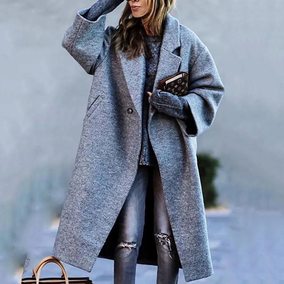 Femme portant un manteau gris oversize, tenant un livre, avec un sac à main en cuir marron. Mode automne-hiver, style décontracté chic.
