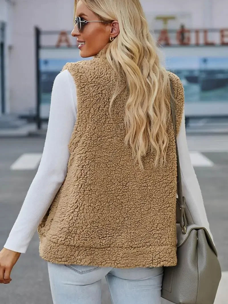 Femme blonde portant un gilet en fausse fourrure beige, manches longues blanches, sac à main gris, lunettes de soleil, rue urbaine en arrière-plan.