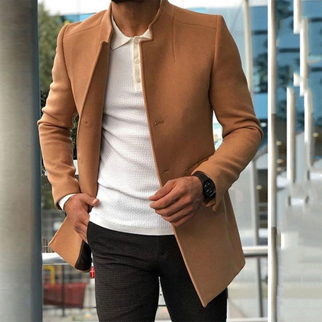 Homme portant un manteau camel élégant, chemise blanche et pantalon foncé, style urbain moderne, montre noire, devant un bâtiment vitré.