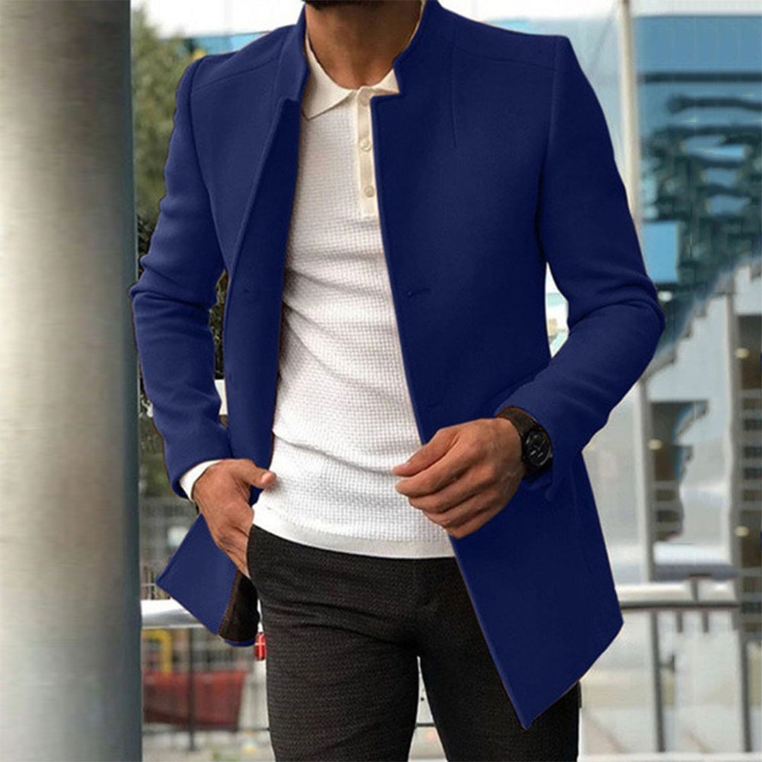 Homme portant un manteau bleu élégant, chemise blanche et pantalon noir, debout à l'extérieur. Mode masculine, style urbain chic.