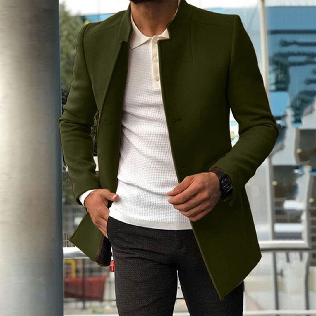 Homme portant un manteau vert élégant, chemise blanche et pantalon gris, posant en extérieur urbain. Mode masculine, style moderne, tenue chic.