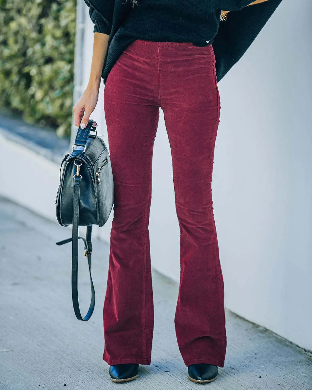 Pantalon patte d'éléphant bordeaux en velours côtelé, femme tenant un sac à main noir, mode automne, style rétro chic, rue pavée.