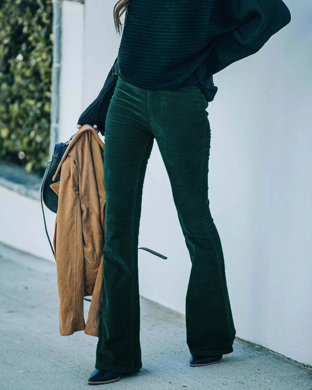 Femme portant un pantalon évasé vert foncé et un pull noir, tenant un manteau marron et un sac à main noir, mode automne-hiver élégante.