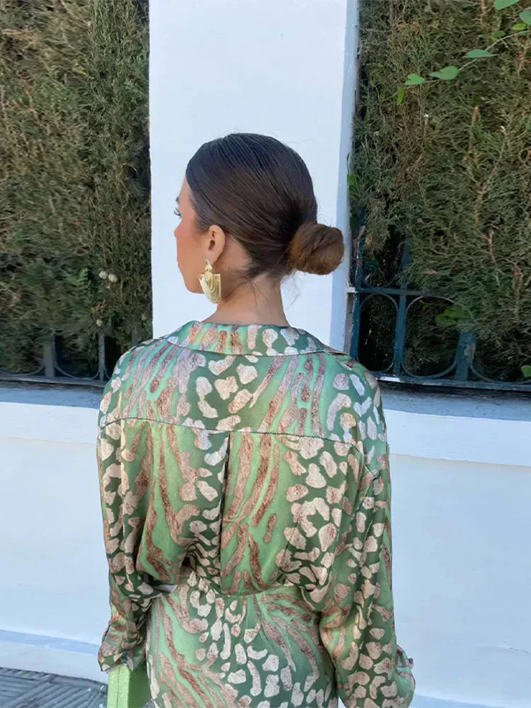 Femme de dos portant une robe verte à motifs léopard, cheveux attachés en chignon, boucles d'oreilles dorées, devant une haie verte et un mur blanc.