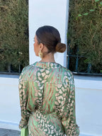 Femme de dos portant une robe verte à motifs léopard, cheveux attachés en chignon, boucles d'oreilles dorées, devant une haie verte et un mur blanc.