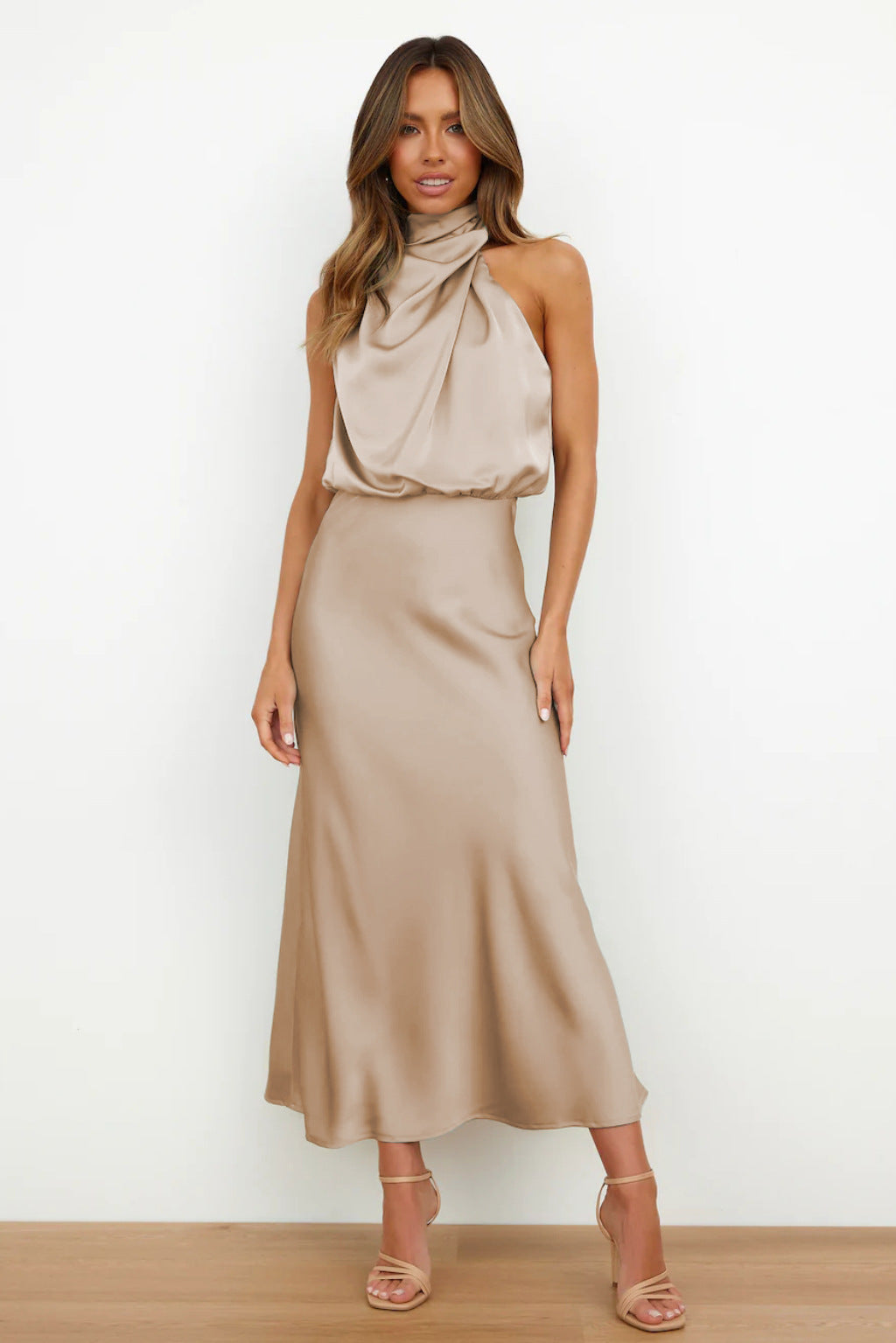 Robe longue en satin beige, sans manches, col drapé, portée par une femme sur fond blanc. Mode élégante, tendance, tenue de soirée chic.