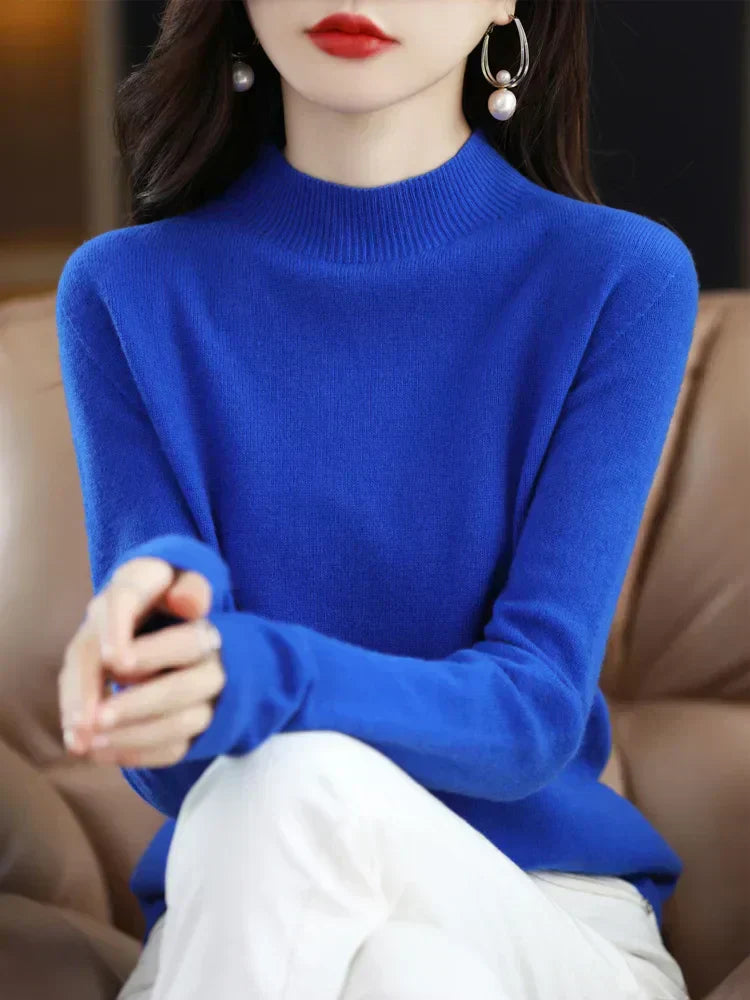 Femme portant un pull bleu vif, assise sur un canapé beige, avec des boucles d'oreilles pendantes et un rouge à lèvres rouge. Mode élégante et moderne.