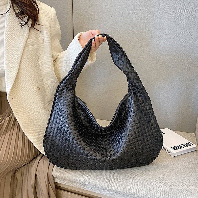 Sac à main noir tressé en cuir, porté par une femme en veste blanche et jupe plissée beige, posé sur un canapé gris. Accessoire mode élégant.