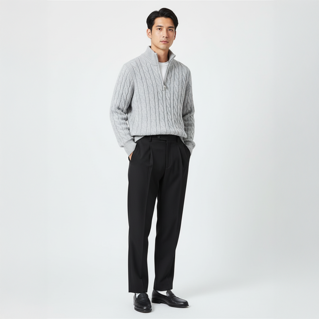 Homme portant un pull gris à col zippé et un pantalon noir, posant debout sur fond blanc. Mode masculine élégante et moderne.