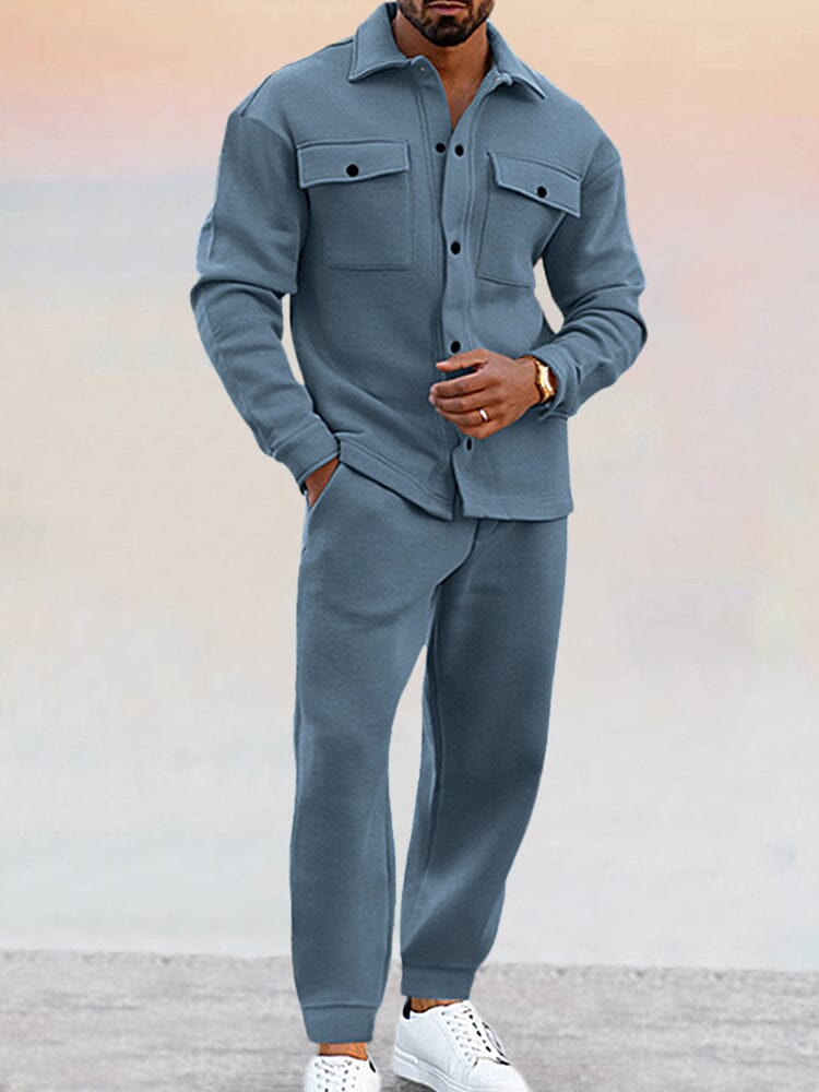 Homme portant un ensemble bleu décontracté, chemise à poches et pantalon assorti, chaussures blanches. Mode masculine élégante et moderne.