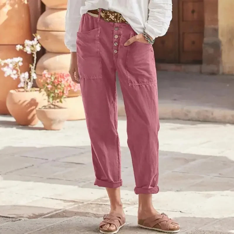 Pantalon rose taille haute avec boutons, porté par une personne en chemise blanche, sandales marron, fond de pots en terre cuite et fleurs.