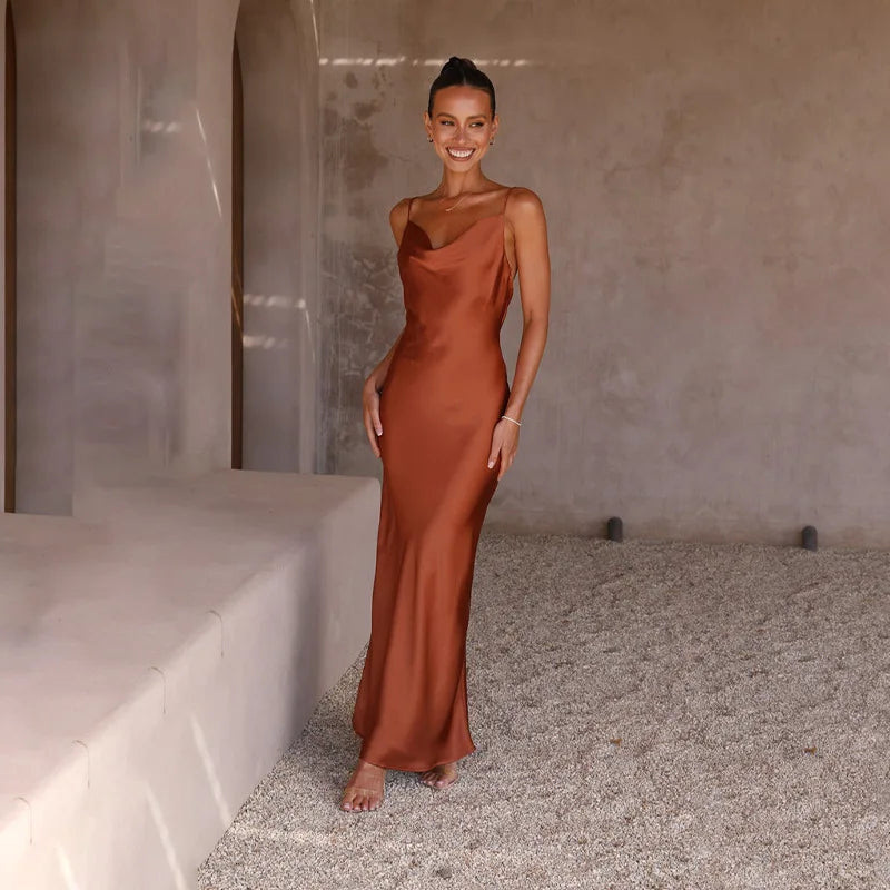 Femme souriante en robe longue satinée marron, posant pieds nus sur un sol en gravier, devant un mur beige. Mode élégante, tenue soirée chic.