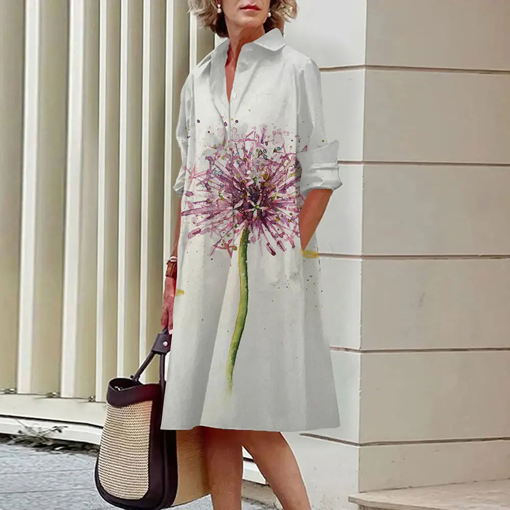 Robe blanche à manches longues avec motif floral coloré, femme tenant un sac à main beige et noir, devant un mur moderne. Mode élégante et décontractée.