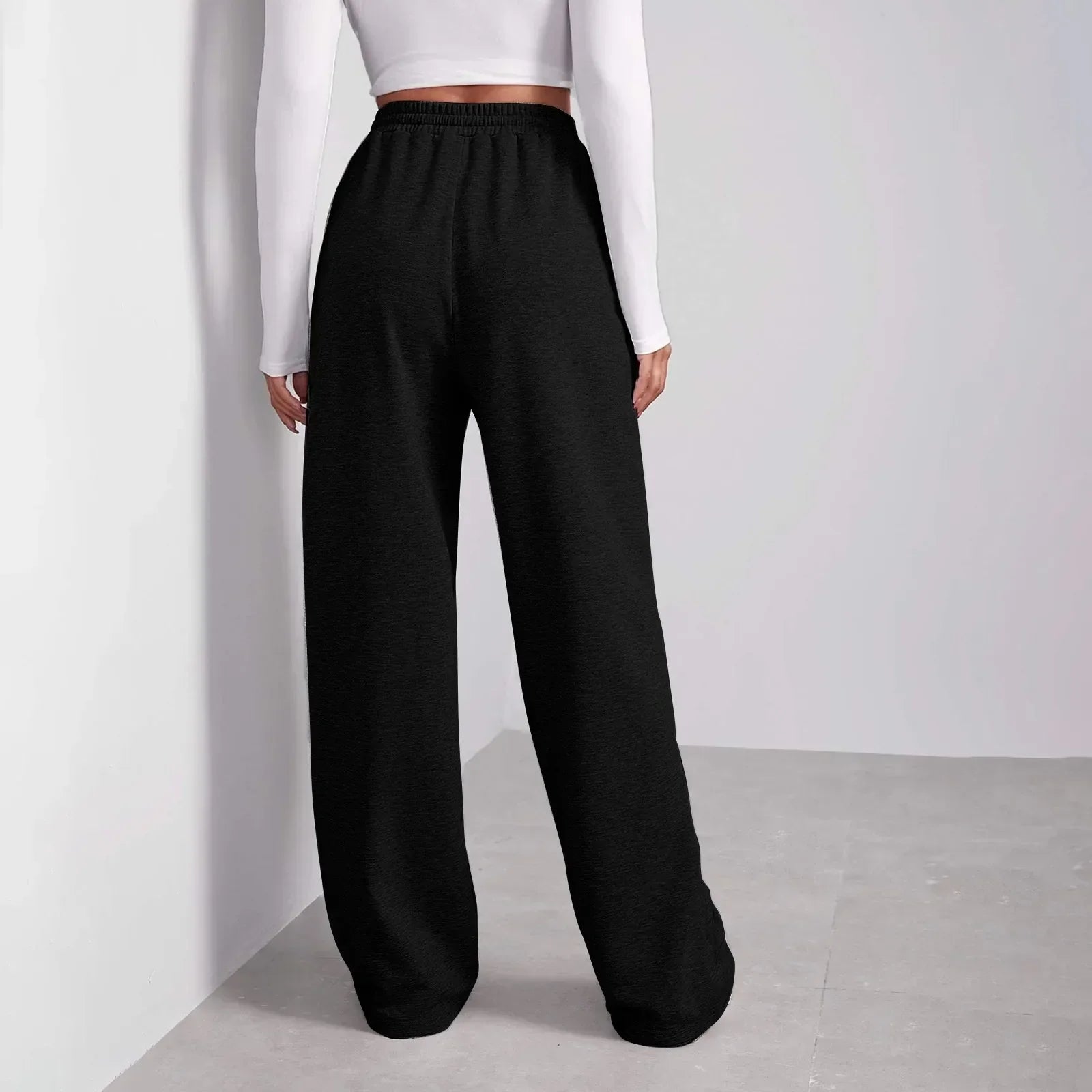 Femme portant un pantalon large noir taille haute, vue de dos, avec un haut blanc à manches longues. Mode décontractée, tendance automne-hiver.