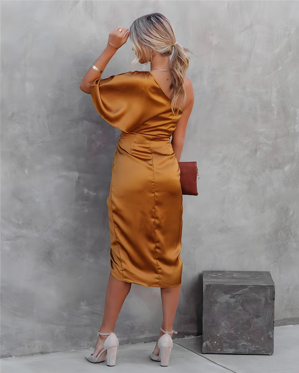 Femme en robe satinée dorée, dos à la caméra, tenant une pochette marron, posant contre un mur gris. Mode élégante, tenue de soirée chic.
