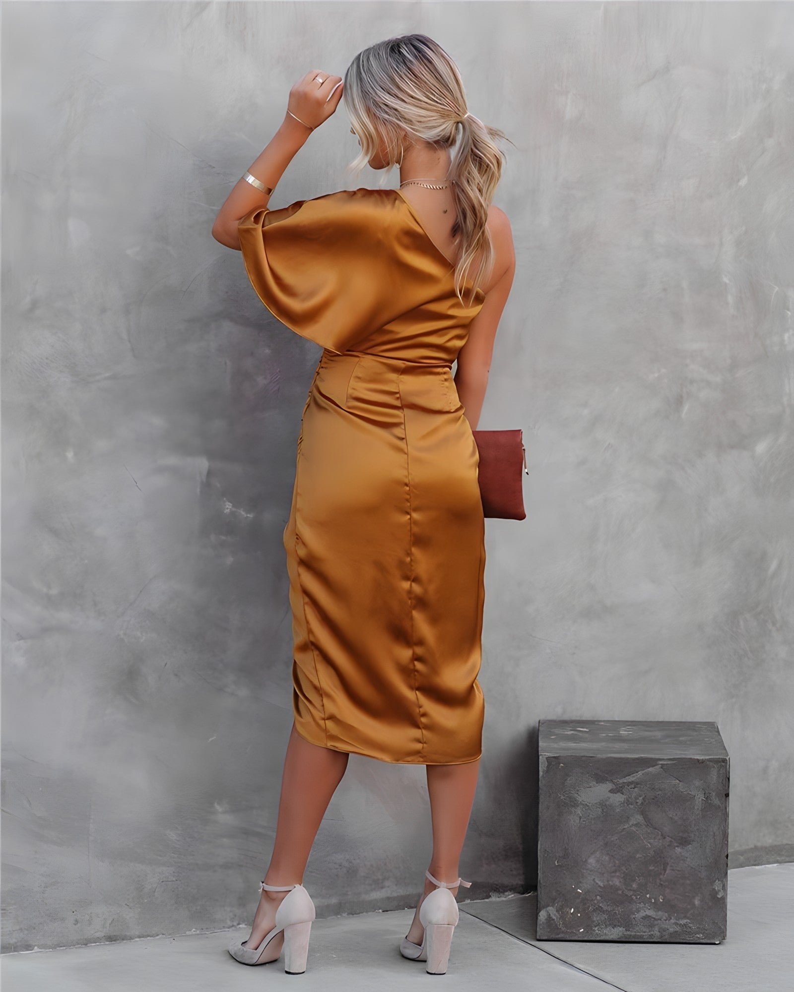 Femme en robe satinée dorée, dos à la caméra, tenant une pochette marron, posant contre un mur gris. Mode élégante, tenue de soirée chic.