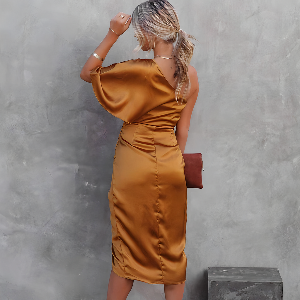 Femme en robe satinée dorée, dos tourné, tenant une pochette marron, posant devant un mur gris texturé. Mode élégante, tenue soirée chic.