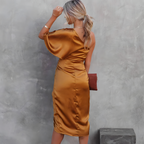 Femme en robe satinée dorée, dos tourné, tenant une pochette marron, posant devant un mur gris texturé. Mode élégante, tenue soirée chic.