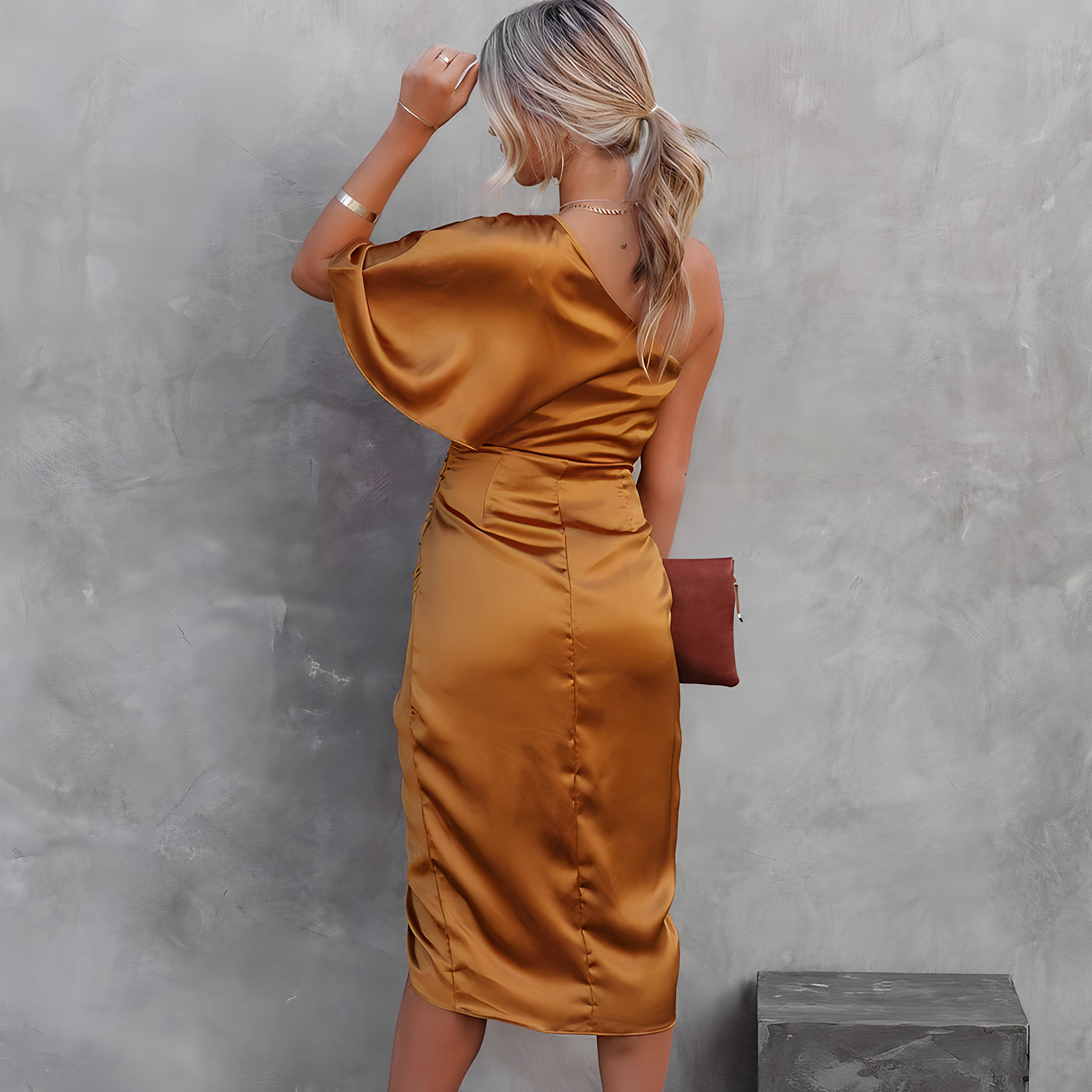 Femme en robe satinée dorée, dos tourné, tenant une pochette marron, posant devant un mur gris texturé. Mode élégante, tenue soirée chic.