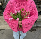 Femme en pull rose vif tricoté, tenant un bouquet de tulipes roses et de gypsophiles, debout sur un trottoir urbain. Mode hiver, style décontracté.