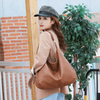 Femme portant un sac à main en cuir marron, vêtue d'un blazer beige et d'une casquette à carreaux, posant devant un mur de briques et une plante verte.
