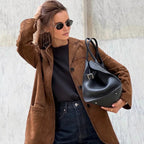 Femme élégante portant un manteau marron, lunettes de soleil rondes et sac à main noir en cuir, posant devant un mur en marbre.