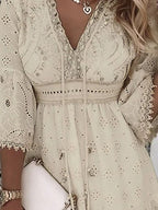 Robe bohème chic en dentelle beige avec manches longues évasées, détails brodés et col en V, accessoirisée d'un collier et d'une pochette blanche.