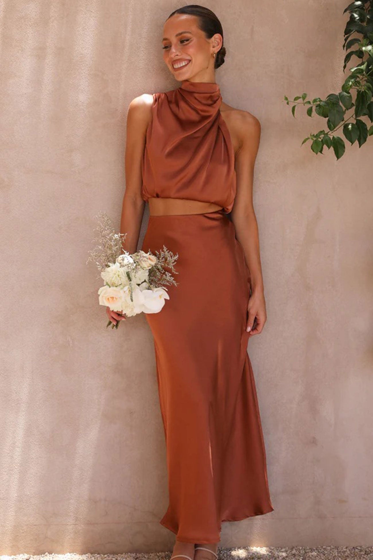 Femme en robe longue marron élégante, tenant un bouquet de fleurs blanches et roses, posant contre un mur beige avec une plante verte.