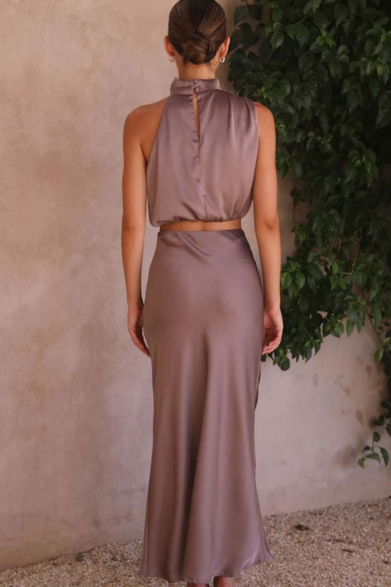 Robe longue en satin taupe, dos nu, élégante et fluide, portée par une femme, devant un mur texturé avec feuillage vert.