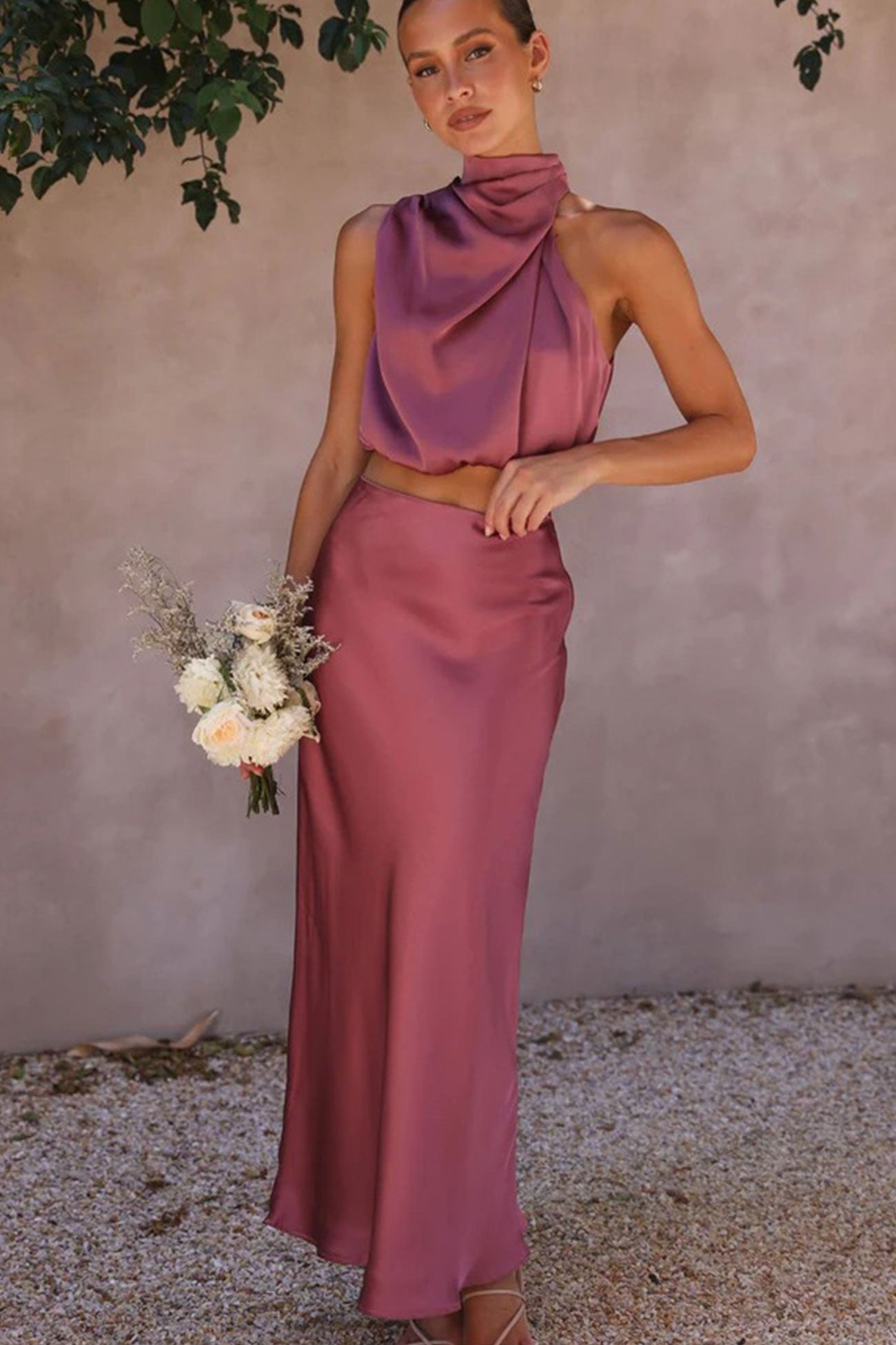 Femme en robe longue satinée rose, tenant un bouquet de fleurs blanches et séchées, posant devant un mur beige. Mode élégante, tenue de soirée.