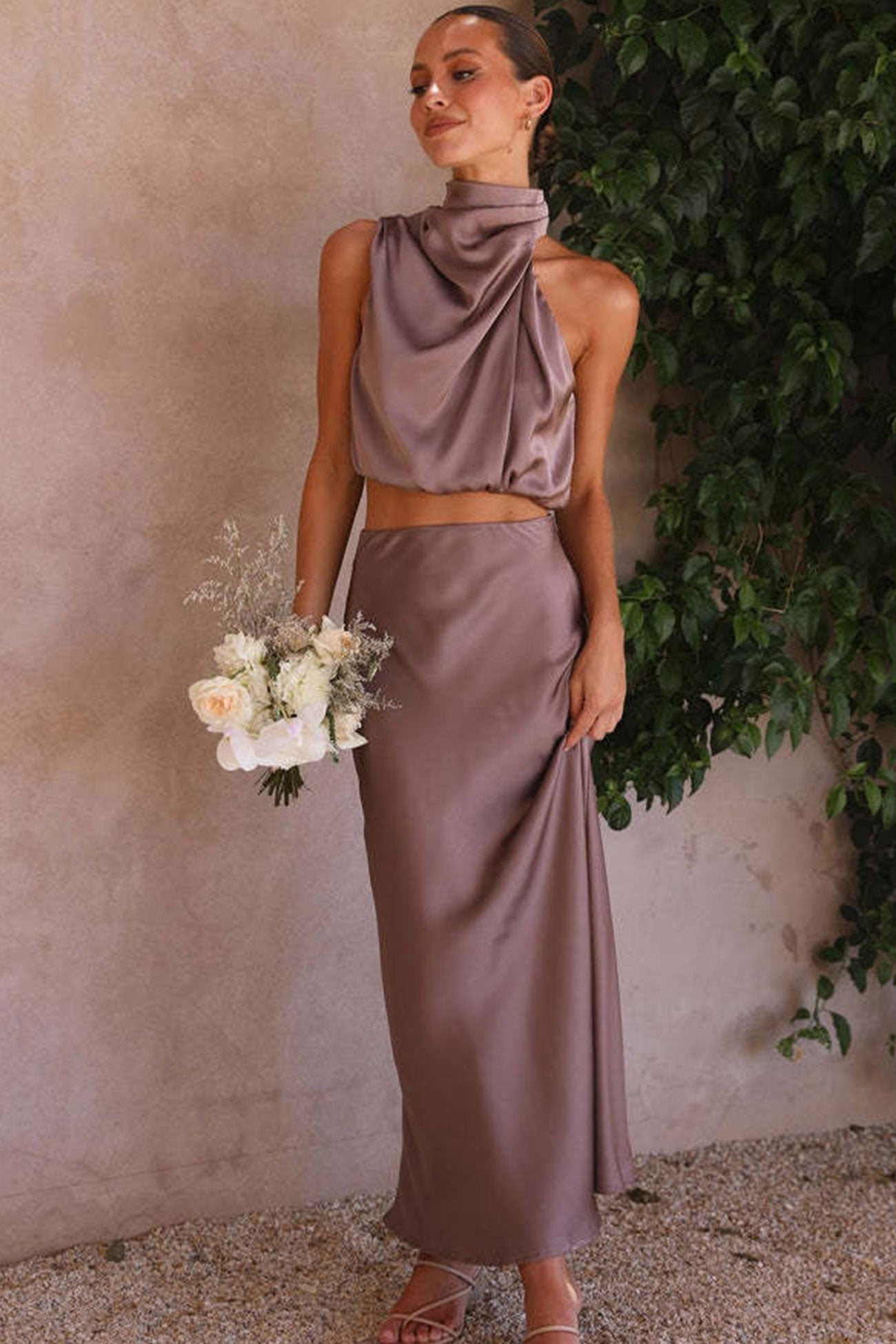 Femme en robe de soirée satinée taupe, tenant un bouquet de fleurs blanches, posant devant un mur texturé avec feuillage. Mode élégante et chic.