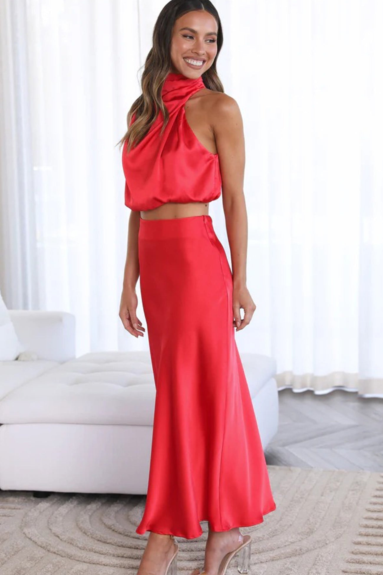 Femme portant une robe longue rouge en satin, sans manches, avec un col haut, posant dans un salon moderne. Mode élégante et tendance.