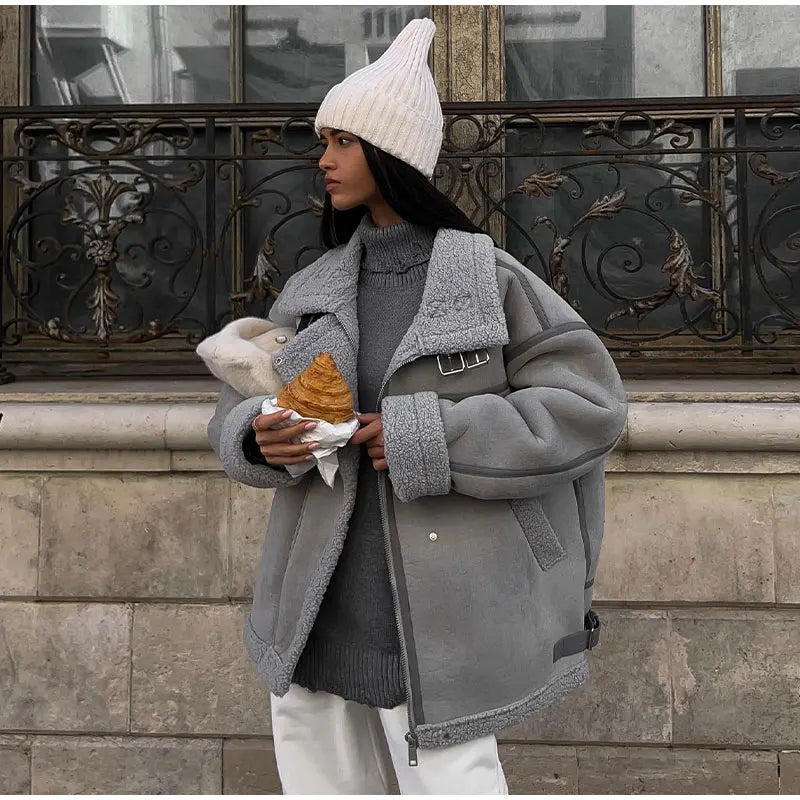 Femme en manteau gris et bonnet blanc tenant un croissant, devant une fenêtre ornée. Mode hiver, style urbain, tenue décontractée.