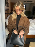 Jeune femme blonde portant un manteau marron et des lunettes roses, tenant un sac à main noir élégant dans une chambre moderne.