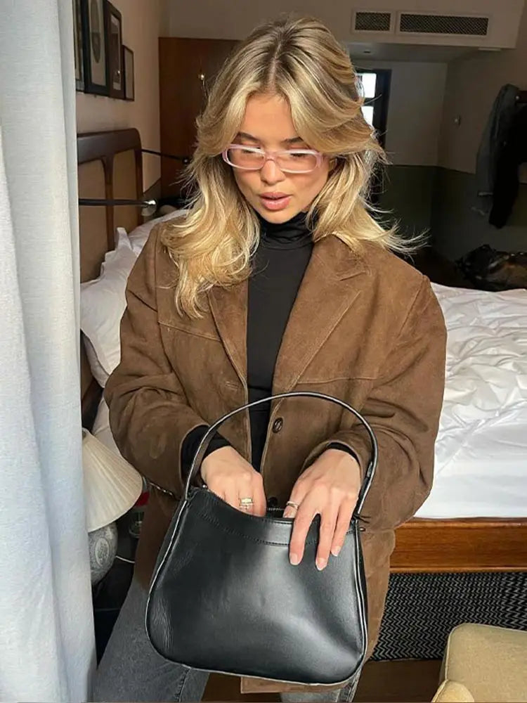 Jeune femme blonde portant un manteau marron et des lunettes roses, tenant un sac à main noir élégant dans une chambre moderne.