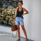 Femme en débardeur noir et short en jean, portant des sandales et un sac à main marron, posant contre un mur blanc. Mode estivale décontractée.
