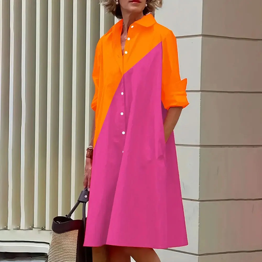 Robe chemise color-block orange et rose, manches longues, portée par une femme tenant un sac noir, devant des stores verticaux blancs.