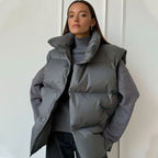 Femme portant une doudoune grise sans manches sur un pull gris, posant devant un mur blanc. Mode hiver, style décontracté, tendance vestimentaire.